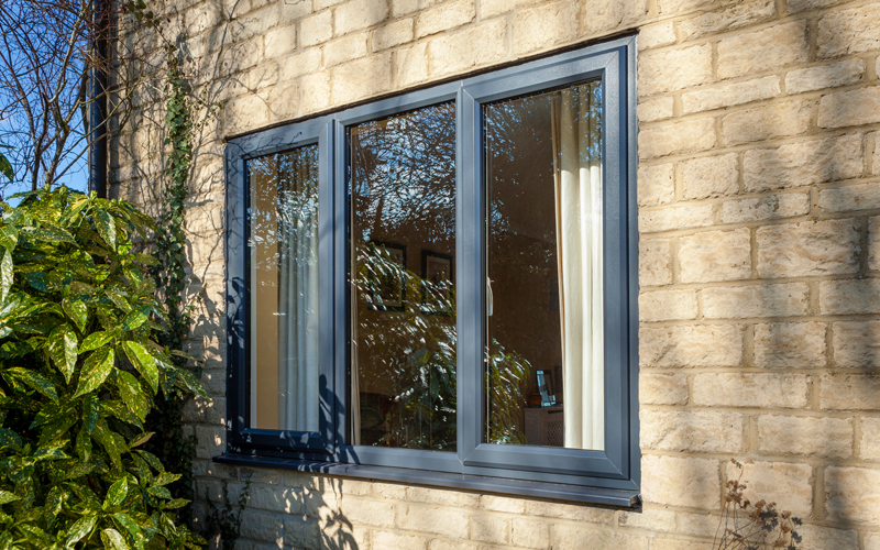 Exterior view of a building with a three-section uPVC casement window featuring a dark frame, installed in a sandstone wall. Reflective glass panes show a glimpse of the interior, while lush green foliage hugs the wall's corner, adding a touch of nature to the scene.