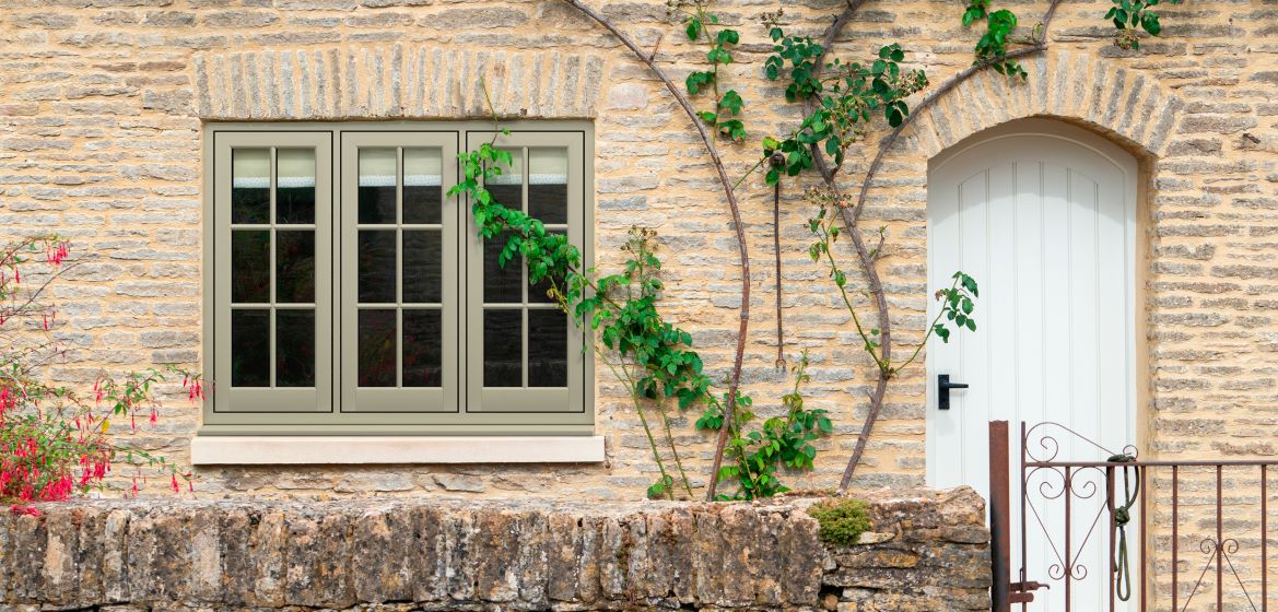 Exterior view of a stone cottage with a taupe double window framed by climbing green ivy on the left and an arched white door on the right. In front of the rustic stone wall, there is a low stone fence with pinkish-red flowers draping over it, and a simple wrought-iron gate partially open, inviting entrance to the charming home